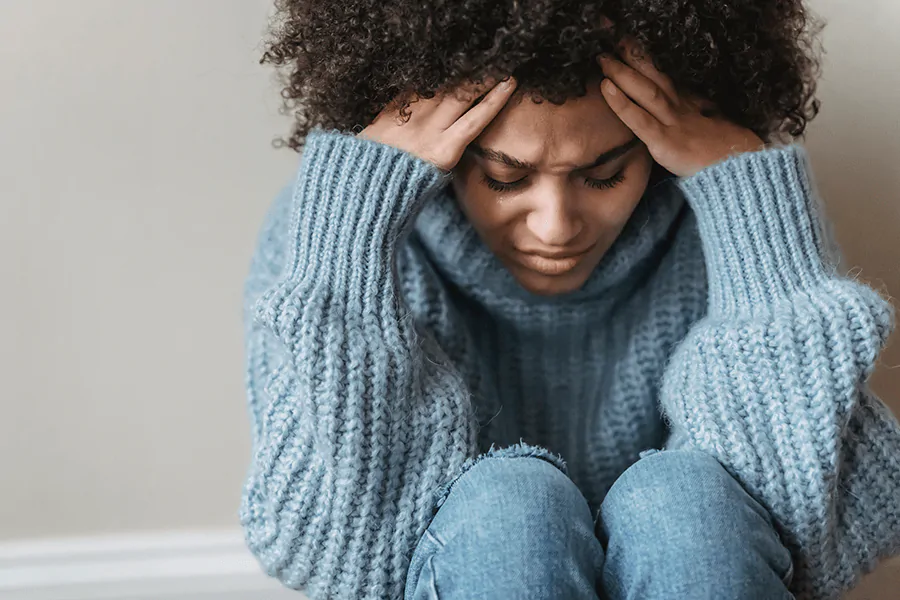 Mulher negra sentada, de olhos fechados, olhando para baixo, com a mão na testa, passando por momento de preocupação constante em busca de uma solução que pode ser a terapia online com uma psicóloga qualificada como a Laís Reis.